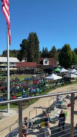 The video opens with a close-up of an American flag against a clear blue sky. It then pans to a grassy area filled with people sitting in numerous colorful lawn chairs. Some people are standing and walking around the chairs. In the background, there are several buildings, including one with a red roof and white trim, and a larger building with a white facade. Tents are set up on the grass. Further in the distance, a body of water with several boats docked is visible, surrounded by a dense forest. The camera then moves to show a wooden pier with people sitting at picnic tables and walking. More boats are seen on the water, and a few kayaks are being paddled. The video ends with a view of the water, the boats, and the tree-covered shoreline.
