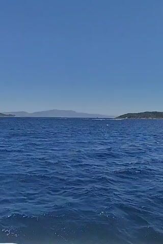 A boat is moving across a large expanse of blue water under a clear blue sky. Islands with green, tree-covered hills are visible in the distance, extending across the horizon. The water has gentle waves. In the middle distance, a white boat with a few visible passengers is moving across the water. The video appears to be taken from another boat, as the edge of a white railing is visible in the bottom foreground.