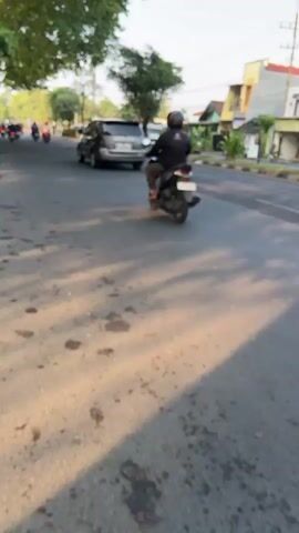 The video shows a street scene in Kludan, East Java. The time of day appears to be late afternoon, with long shadows cast across the asphalt road by trees. The weather seems clear and sunny.

The main subjects are several motorcycles and cars traveling along the road. One motorcycle with a rider wearing a dark jacket and helmet is prominently featured in the foreground, moving away from the camera. Another motorcycle, also with a rider, is visible further down the road. Several cars are also seen, including a dark SUV to the right and other vehicles in the distance. The background shows residential buildings, suggesting a populated area. Trees line the left side of the road, providing some shade.

The activity taking place is the movement of vehicles along a public road. There are no discernible emotions expressed by the individuals due to the distance and lack of clear facial views.

There is no visible text in the media file.