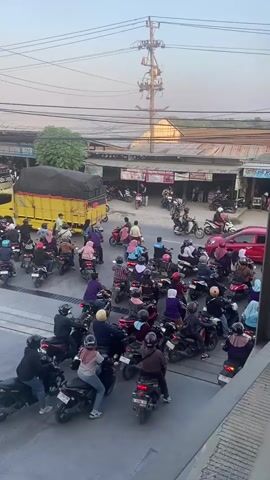The video shows a congested street scene with numerous motorcycles and a few cars. People are seen riding motorcycles, waiting in traffic, and interacting in small groups. The environment appears to be a commercial area with shops and buildings lining the street, and overhead power lines are visible. The overall impression is of a busy urban or semi-urban setting experiencing traffic congestion.