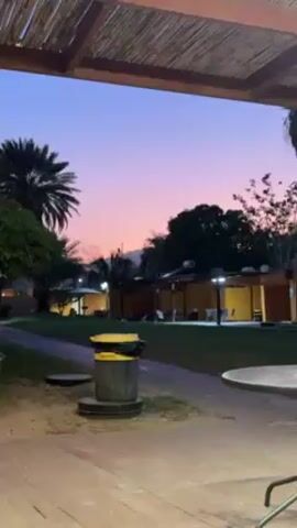 Okay, here's a detailed description of the scene in the video:

**Setting:** The video appears to be shot in an outdoor area, possibly a resort or park, with a relaxed and peaceful atmosphere. It is evening, as indicated by the twilight sky.

**Environment:** The foreground shows a partially covered patio area with a table and chairs. In the middle ground, there's a grassy area with scattered trees, palm trees, and small buildings or huts. The sky in the background displays a gradient of colors, transitioning from a deep purple at the top to a pinkish-orange near the horizon. A mountain range is faintly visible in the distance.

**Objects:** Key objects include:
- A trash can in the foreground, near the patio area.
- Tables and chairs on the patio.
- Small buildings or huts with lights on inside.
- Trees, including palm trees.

**People:** Only one person can be barely seen, walking in the distance.

**Actions:** The video is static.

**Overall Impression:** The scene evokes a sense of tranquility and peacefulness. The focus is on the natural beauty of the sunset and the calm environment.