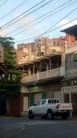 The video shows a street scene with several buildings, a white Toyota pickup truck, and a white sedan. The buildings appear to be a mix of old and new construction, with some sections appearing unfinished. There are also a lot of overhead wires visible. The pickup truck is parked on the street, and the sedan is driving past it. The video is shot from a low angle, possibly from a moving vehicle. The lighting suggests it is late afternoon or early evening.
