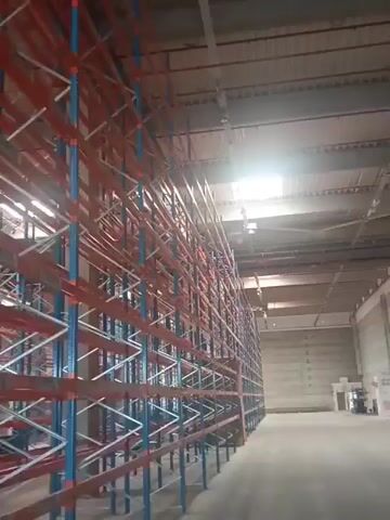 The video shows a long shot looking up at a large industrial shelving unit inside a warehouse. The shelving unit is tall and appears to be made of metal. It is painted in blue and orange. The shelves are empty. The lighting suggests it is daytime, with sunlight streaming in from above. The overall environment is clean and orderly.