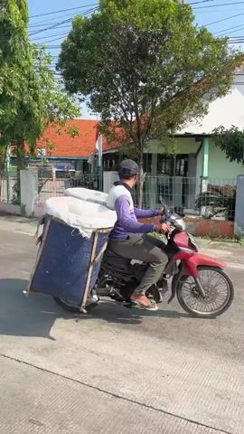A person wearing a purple and white shirt, dark pants, and a black cap rides a red motorcycle on a paved road. The motorcycle has a large blue bin attached to the back, covered with a white tarp, and appears to be filled with white items. The person is looking forward and steering the motorcycle. The environment is an outdoor setting with a building in the background, trees, and a sidewalk. The sun is shining, casting shadows.