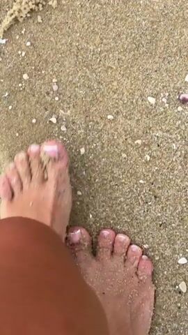 The video shows a person's foot walking on a sandy beach. The foot is bare and has painted toenails. An anklet with beads is visible on the ankle. The sand is light brown and appears dry, with some small shells and debris scattered on it. The camera perspective is from above, looking down at the foot and the sand. The video is short and focuses on the act of walking.