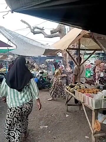The video pans across an outdoor market stall. People are browsing and shopping for produce. The stall features a variety of fresh vegetables, including cucumbers, eggplants, green onions, and corn. Several individuals, predominantly women wearing headscarves, are seen examining the produce or interacting with vendors. One vendor is grilling food on a portable grill. The market appears to be set up under makeshift shelters with tarps and awnings providing shade. The ground is dirt and gravel. The overall atmosphere suggests a busy, open-air marketplace.