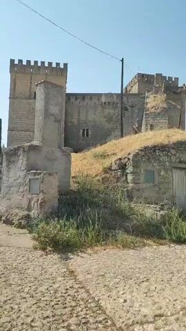 The video pans across an arid, historical landscape under a clear blue sky. The scene features stone structures, some appearing to be ancient dwellings partially built into hillsides, and others resembling parts of a castle or fortress with crenellated walls visible in the distance. The ground is a mix of rough cobblestone and dry, sparse vegetation. Wooden doors and small windows are visible on the stone buildings. The overall impression is one of a preserved historical site in a dry, sunny climate. No people or animals are visible in the video.