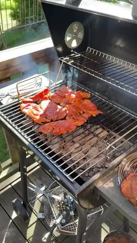 A close-up, eye-level shot shows marinated meat cooking on a charcoal grill. The meat, a vibrant red and orange color, is arranged in a single layer across the grill grates. Wisps of smoke rise from the grill, indicating it is hot and actively cooking the food. The grill itself is black and sits on a wooden deck. In the background, a railing and greenery are visible, suggesting an outdoor setting, likely a backyard or patio. Sunlight streams in from the right side, casting bright highlights on the grill and meat, suggesting it is daytime, possibly late morning or afternoon, with fair weather. The overall mood is one of casual outdoor cooking, perhaps a barbecue or a casual meal being prepared. No visible text is present in the media.