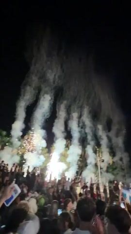 A large crowd of people are gathered outdoors at night, watching a fireworks display. Many in the crowd are holding up their phones to record the event. The fireworks are creating a large amount of smoke and bright lights, obscuring the sky and some of the background scenery. Some trees are visible in the background, illuminated by the fireworks. The overall impression is one of excitement and celebration.