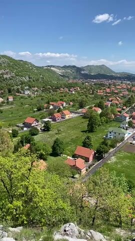 The video shows an overview of a rural town or village, set against a backdrop of green hills and a bright blue sky dotted with fluffy white clouds. The village is characterized by houses with terracotta tile roofs, scattered across a landscape of green fields and trees. A few roads meander through the community. The foreground features rocky terrain with trees, adding a sense of height and perspective to the shot. The overall atmosphere is calm and serene.