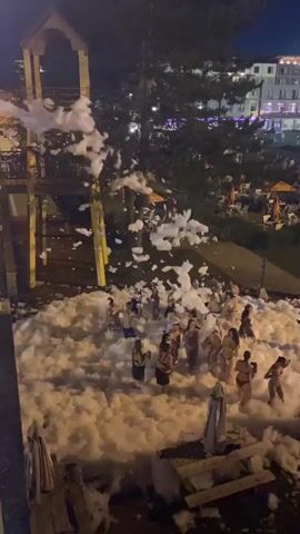 The video opens on a pile of white foam being sprayed from an elevated platform, which is partially visible at the top left of the frame. Below this platform, numerous people are gathered in the foam, some appearing to be playing. The setting appears to be an outdoor venue at night, with trees and buildings visible in the background. Further in the background, there is a large building with lights, possibly a resort or hotel, and a Ferris wheel is visible with colorful lights. The ground is covered with grass, and picnic tables with umbrellas are scattered throughout the area. Some people are standing around the perimeter of the foam area, while others are interacting within the foam. The overall atmosphere seems to be festive, with the foam party activity being the central focus.