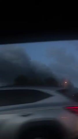 The video shows a view from inside a moving vehicle, likely a car, looking out the passenger side window. The sky is dark and filled with ominous, swirling clouds, suggesting an approaching storm or severe weather. The foreground consists of the vehicle's window frame and the side mirror, which are partially visible. The road outside is wet and reflecting light. Trees and a bridge are partially visible against the backdrop of the stormy sky, adding to the overall gloomy atmosphere.