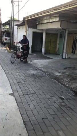 A person wearing a dark headscarf and a dark patterned dress is walking a bicycle on a paved street. The bicycle has a basket on the front. The street is made of dark bricks with a white border. In the background, there are buildings with yellow trim and garage doors. A car is parked in the distance.