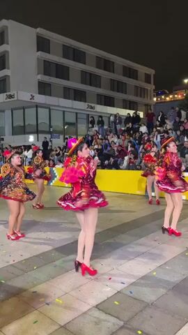 The video shows a group of women in brightly colored, sequined costumes performing a dance outdoors at night. They are wearing short, flared dresses, matching hats, and high-heeled shoes. Their costumes are ornate with intricate patterns and embellishments. The dancers are moving in unison, with their arms outstretched and their skirts swirling. The background shows a crowd of spectators gathered behind a yellow barrier, and a modern building with large windows. The ground is paved and scattered with colorful confetti. The lighting suggests a festive atmosphere, possibly a parade or cultural event.
