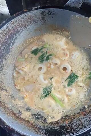 A close-up overhead shot shows a person stirring a dish in a wok. The dish appears to be noodles with seafood and vegetables in a broth. The wok is old and seasoned, with a dark, metallic sheen. The noodles are curly and yellow, and the seafood consists of rings of calamari and small meatballs. Green leafy vegetables are also visible. The broth is a creamy beige color and is bubbling, indicating that it is hot. The person is using a metal spatula to stir the ingredients, and the noodles and other components are swirling in the wok. The lighting is natural and appears to be coming from above. The background is blurred, but it seems to be an outdoor setting.