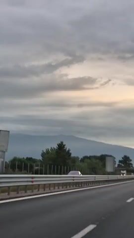 The video shows a view from a vehicle traveling on a highway. The highway has multiple lanes and is separated by a metal barrier. There are cars and trucks on the highway. On the side of the highway, there is a fence and trees. In the background, there are mountains under a cloudy sky. The lighting suggests it is either early morning or late evening.