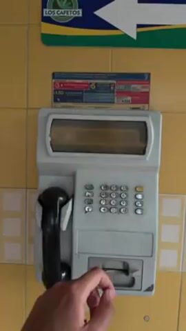 A person's hand is shown dialing a number on a public payphone. The payphone is a light gray color with a black handset. The keypad has standard numbers, and the hand presses the keys one at a time. The phone is mounted on a yellow tile wall. Above the phone is a sign with call rates and an arrow pointing left with the words "Los Cafetos." The video focuses on the phone and the hand interacting with it, with no other people visible.