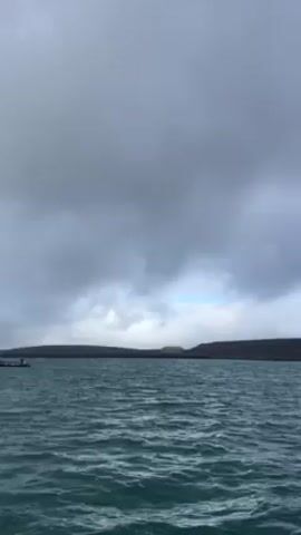 The video shows a view from a boat on choppy turquoise waters. The horizon line is dark land with a few patches of snow on the right side and some buildings visible on the left. In the distance, large rusted metal tanks are visible. The sky is overcast with grey clouds. There are no people visible in the video.