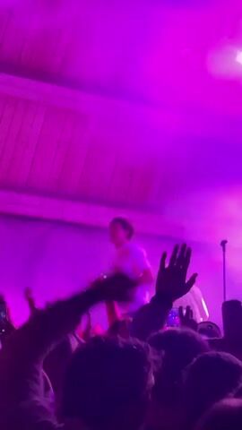 A crowded concert takes place on a stage with a large, curved wooden structure above. A performer wearing a light-colored t-shirt stands at the front of the stage, holding a microphone to their mouth and appearing to sing. Pink and purple stage lights illuminate the scene, creating a vibrant atmosphere. In the foreground, a diverse crowd of people can be seen with their hands raised in the air, some holding up their mobile phones to record the performance. The overall impression is of an energetic and engaging live music event.