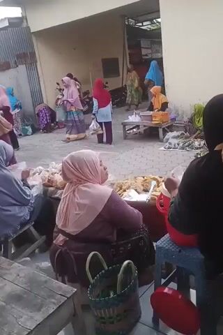 The video shows a marketplace scene with several women dressed in hijabs and traditional attire. They appear to be vendors and customers. Some women are seated behind stalls laden with goods, possibly food items like bananas and other produce. Others are standing and interacting, suggesting a bustling atmosphere. The environment appears to be an outdoor or semi-outdoor market, possibly with stalls set up in an alley or courtyard with buildings on either side. There are also some motorcycles parked in the background, indicating that transportation is present. The lighting suggests it is daytime.