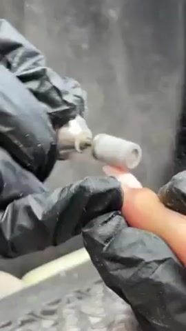In this close-up shot, a person wearing black gloves is using a small, cylindrical grinding tool to work on a fingernail. The tool is attached to a handheld device. The person appears to be carefully shaping or smoothing the surface of the nail. The background consists of a greyish, somewhat indistinct surface, possibly a workbench or table with scattered debris.
