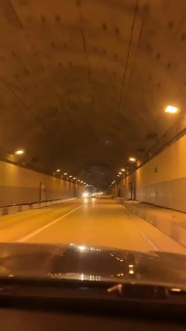 The video shows a car driving through a tunnel. The tunnel has a curved ceiling and is lit by bright lights. Another vehicle passes in the opposite direction. There are large, round vents in the ceiling. The windshield of the car is dusty.