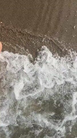 The video captures a close-up, overhead view of a person's feet wading in shallow, murky water, likely at a beach or shore. The water is disturbed by the movement of the feet, creating foamy white waves and ripples that cascade over the skin. The bottom appears to be sandy or muddy. The lighting suggests it is daytime.
