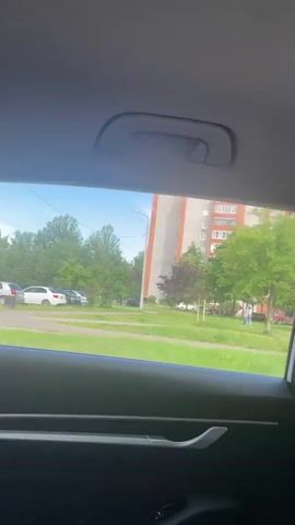 The video captures a view from inside a vehicle, looking out the rear passenger window. The scene outside is an urban residential area with a bright, sunny sky. In the foreground, the interior of the car is visible, including a portion of the door panel and a handle. Beyond the window, a grassy area is present, with trees and a tall, multi-story apartment building in the background. Several people, appearing to be a mix of adults and possibly young adults or teenagers, are gathered on the grass. They are standing in a loose group, some interacting with each other, while others appear to be observing something. One individual is walking away from the group towards the right side of the frame. The overall impression is of a casual outdoor gathering or event taking place in a typical urban setting.