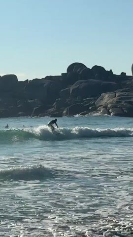 The video shows a person in a wetsuit surfing in the ocean. The person is seen from the back, riding a small wave toward the shore. In the background, there are large, dark-colored rock formations. The sky is blue, and the water is a mix of blue and white due to the waves. Another person can be seen in the distance to the left of the surfer. The camera remains mostly static throughout the video.