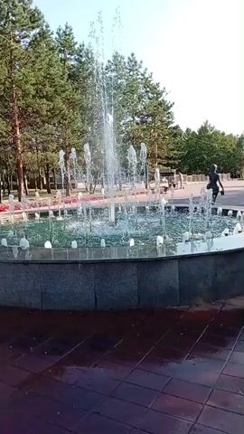 A fountain sprays water in a park. Trees line the background. Several people are walking around the fountain. One person is in the fountain's water. The ground around the fountain is wet.