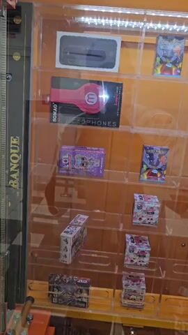 This is a close-up, medium-shot, vertical video of a claw machine filled with various toys and small electronic items. The machine has multiple shelves displaying the prizes. On the top shelf, there is a black smartphone-like object and a colorful box. Below that, there is a pink pair of headphones in packaging. The next shelf down has a purple box with cartoon characters and a smaller box with a similar design. To the right, there is a box with a Star Wars theme and another small box. The shelves below are filled with more small boxes, some appearing to be blind bags or collectible toy sets, and a few small plastic figurines. The bottom section of the machine shows the claw mechanism and a coin slot with pricing information displayed in euros. The machine has a "BANQUE" logo visible on the side. The lighting is bright, illuminating the prizes inside the glass display case.