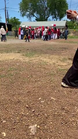A low-angle, eye-level shot shows a gravelly, dry patch of ground in the foreground with sparse green grass. A person in black pants and a white shirt, wearing a red headscarf, walks away from the camera towards the right. Their feet are visible, one of which is a white sneaker with a black sole. In the middle ground, a group of people are gathered under a white tent. Red and white balloons are tied to the tent. The people are of various ages and are dressed in a mix of casual and traditional clothing, with many wearing red. Some individuals are holding items, and one person in the foreground to the left is crouched down, possibly interacting with something on the ground. The background features a large tree and a bright sky with some clouds. The overall impression is of an outdoor gathering or event.