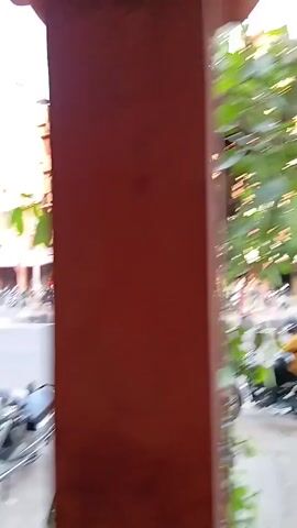 The video shows a view from an enclosed space, likely a shop or building, looking out onto a street in India. Across the street stands a large, multi-story building painted in a reddish-pink hue. The building has rows of windows and what appear to be decorative arches along the ground floor. Parked motorcycles line the sidewalk in front of the building. A person wearing dark clothing is walking across the street, holding what might be a cell phone. An auto rickshaw, also known as a tuk-tuk, passes by in the background. The foreground is slightly obscured by a plant and some other items inside the building from which the video is being taken.