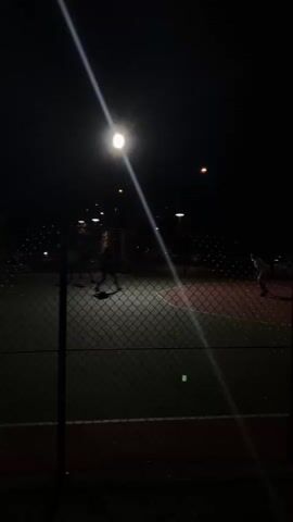 The video shows a night-time scene on an outdoor sports court, likely a basketball or tennis court, illuminated by a single bright overhead light. Several people, appearing as silhouettes, are visible on the court, engaged in some activity, possibly a game or practice. A chain-link fence runs along the foreground, partially obscuring the view of the court. The background is dark, with a few other distant lights visible, suggesting a suburban or park setting. The camera is static, and the video is short, capturing a brief moment of activity.