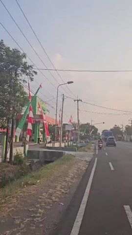 The video shows a street scene during daylight. There are buildings on the left side of the road decorated with red and white flags. Power lines and utility poles are visible throughout the scene. Traffic, including cars and motorcycles, moves along the asphalt road. The sky is clear with some light clouds. The sun is visible in the distance, casting a warm glow. The overall environment appears to be an urban or semi-urban setting.
