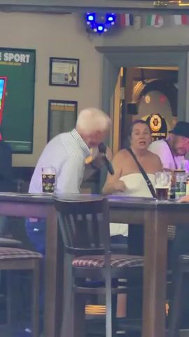 The video shows a group of people sitting around a table in what appears to be a pub or restaurant. An older man with white hair, wearing a striped shirt, is holding a microphone and singing. A woman in a white top stands next to him, gesturing with her hand. A man in a hat is seated behind them, watching the performance. There are glasses of beer, a bottle, and other items on the table. Decorations and signs are visible on the walls in the background. The camera moves slightly, focusing on the older man as he sings. The lighting appears to be dim and ambient.
