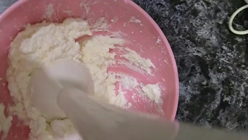 A close-up shot shows a pink bowl containing a white, creamy substance that appears to be cottage cheese or ricotta cheese. A white immersion blender is being used to blend the cheese. The blender is submerged in the cheese and is actively mixing it. The background is a dark, speckled countertop. The lighting is overhead and appears to be artificial.