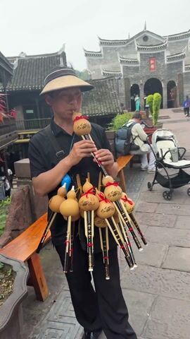 A man wearing a sun hat and a black outfit stands on a brick walkway and plays a musical instrument called a hulusi. He holds a collection of the instruments, all identical in design with a gourd-shaped top tied with a red ribbon, displayed on his body. In the background, there is a traditional Chinese building with gray tiled roofs and a gate, where several people are walking. Other people are sitting on a bench to the side. A stroller is also visible. The setting appears to be an outdoor area with stone walkways, plants in pots, and a wooden bench.