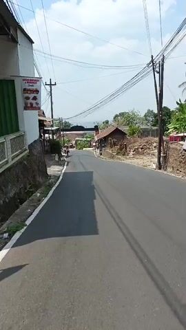 A paved road passes through a rural village with houses on either side. Overhead power lines crisscross the sky. Several children in school uniforms, consisting of white shirts and maroon pants or skirts, are walking along the side of the road. One boy in the foreground is wearing a backpack and walking barefoot. Another group of children further down the road are also heading in the same direction. The weather appears to be sunny with some clouds. The scene suggests a typical day in a village where children are on their way to or from school.