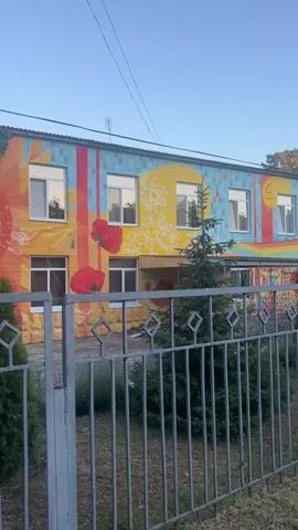 A side-view, medium-long shot shows a building with a vibrant mural on its facade, depicting sunflowers and poppies. The mural features a sun-like design and a cascade of green ivy. A metal fence with diamond-shaped cutouts runs across the foreground. Beyond the fence, a small evergreen tree stands in front of the building. In the background, a crowd of people is gathered in a courtyard area, with trees and some structures visible. The sky is clear and blue, suggesting daylight. The camera is panning slowly to the right.