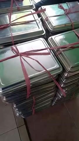 The video shows a person wearing a dark hijab and blue shirt, sitting at a table, and tying red ribbon around stacks of stainless steel trays. The trays are square or rectangular, and some are stacked up to a dozen high. The person is using a red object, possibly a knife or a tool, to cut the ribbon. The environment appears to be indoors, with tiled flooring and a light-colored wall in the background. There are several stacks of these trays, suggesting a large quantity.