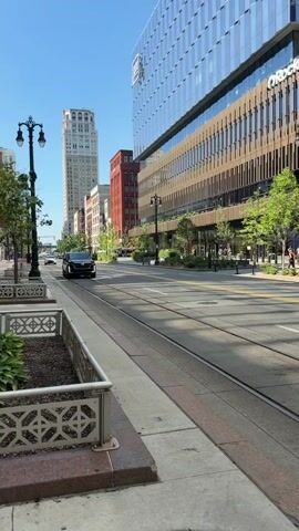 The video shows a black Cadillac Escalade driving on a city street with streetcar tracks. The car is moving towards the camera from the left side of the frame. Tall buildings line the street, one of which has "Rocket" written on it. There are trees and streetlights along the sidewalk. A white car is visible in the background, also moving away from the camera. The sky is blue and clear.