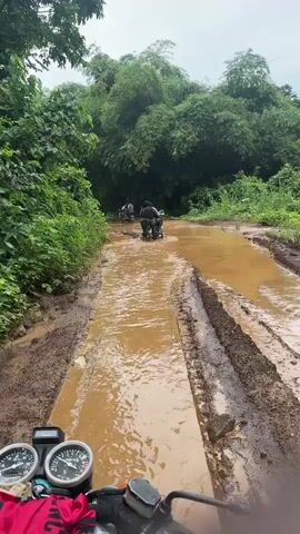 A first-person view from the back of a motorcycle shows a person with short dark hair, wearing a green t-shirt, seated on the motorcycle. The handlebars and instrument panel of the motorcycle are visible in the foreground, with a red garment draped over the tank. The motorcycle is on a deeply rutted, muddy dirt road filled with brown water. Ahead, two other motorcycles are navigating the flooded road, driving through the murky water. The road is surrounded by dense green tropical vegetation, including tall trees and bushes, under an overcast sky.