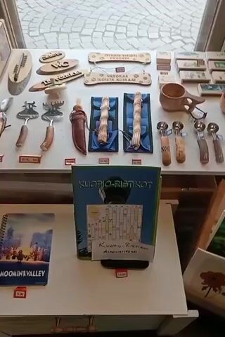 The video shows a retail display of various handicraft items. On a table in the foreground, there are several items including a book with a crossword puzzle, wooden mugs, and carved wooden utensils. To the right, there is a display of greeting cards featuring images of snowmen, the Aurora Borealis, and floral motifs. Further to the right, there is a rack displaying more greeting cards and small decorative items.

In the background, there is a display of crocheted hats in various colors and styles. A wire rack holds many small packaged items, possibly jewelry or accessories. Small Finnish flags are also visible on display. The overall impression is that of a shop selling local crafts and souvenirs. The lighting appears to be natural, coming from a window. The floor in the background appears to be tiled.