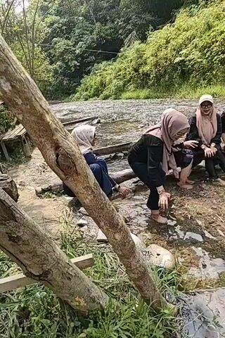 A group of four women, all wearing hijabs, are gathered near a shallow stream in a wooded area. Two of the women are crouched down by the water's edge, with one of them dipping her hand into the stream. The other two women are sitting further back. The environment appears to be natural and somewhat overgrown, with trees and vegetation surrounding the stream. There are some wooden structures and rocks visible in the foreground. The lighting suggests it is daytime.