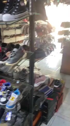 The video shows a store shelf filled with shoes. The shelf is made of metal and has glass shelves. There are many different types of shoes on the shelf, including sneakers, sandals, and dress shoes. The shoes are in a variety of colors and sizes. There are also shoe boxes stacked on the floor at the bottom of the shelf. In the background, more shoes and sandals are hanging from the ceiling and other shelves. The store appears to be well-lit with sunlight streaming in from a window.