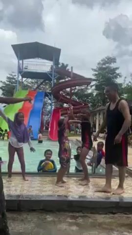 Here is a detailed description of the video:

The video takes place at a waterpark with a large slide structure visible in the background with green, blue, and red slides. Several people, including children and adults, are near the edge of a pool.

People include:
- A girl with a purple hijab.
- Two men, one in a tank top and the other with only an arm visible.
- A girl wearing a dark hijab.
- 3 other children in the pool, including one with a yellow floating toy.

The actions involve a group gathering and seemingly dancing or playfully interacting near the pool's edge, in front of the slide. The atmosphere seems cheerful, with some individuals raising their arms in the air.