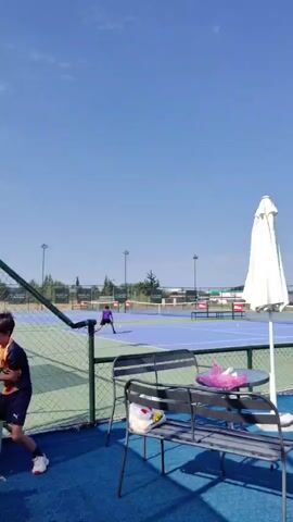 The video shows an outdoor tennis court on a sunny day. A young boy wearing a blue and orange shirt and black shorts is standing on a blue surface, near a green fence. He is holding a tennis racket. In the background, another person in a purple shirt is playing tennis on a blue court. There are two white umbrellas, a metal bench, and a small round table in the foreground. The sky is clear and blue.