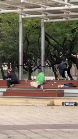A young man attempts a skateboarding trick, falls, and lands on his back. The video starts with a skateboarder in a gray shirt and blue pants standing on a set of brown stairs. He attempts a trick, lands, and immediately loses his balance, landing hard on his back on the light gray and brown tiled pavement. The skateboard slides away from him. A photographer with a red hat and black shirt kneels on the stairs, likely filming the stunt. Another person sits to the right wearing a lime green jacket and khaki pants. Several skateboards and trash are scattered on the pavement. A large modern gazebo-like structure is behind the stairs, framed by green trees.