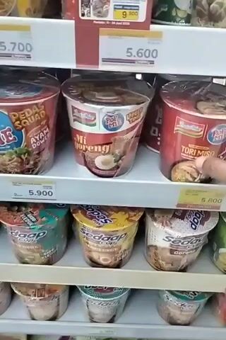 The video shows a close-up of a hand reaching for a cup of instant noodles on a supermarket shelf. The shelf is stocked with various brands and flavors of instant noodles, mostly in cup form. The hand is positioned to grab a red cup labeled "Indomie Pop Mie Tori Miso". The background shows other shelves filled with similar products. The lighting is typical of a supermarket environment. The focus is on the hand and the noodles.